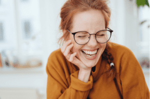 Pretty red-haired girl with pigtail, wearing glasses and orange sweatshirt, laughing